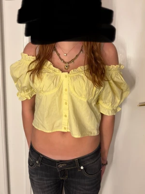 Gul croppad blus med volang - Säljer en ljusgul croppad blus med offshoulder och puffiga korta ärmar. Blusen har knappar framtill och volangdetaljer längs halsringningen och ärmsluten. Perfekt för sommarens alla tillfällen!