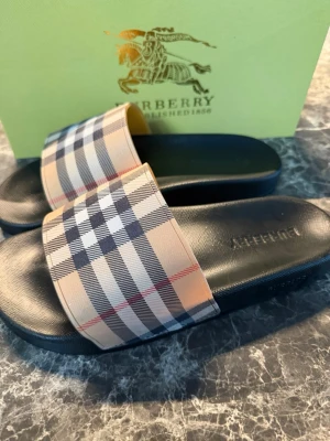 Burberry sandaler med rutig mönster - Säljer ett par snygga Burberry sandaler med klassiskt rutig mönster i beige, svart, vitt och rött. Sandalerna har en bred rem över foten och svart platt sula med Burberry-logga. Perfekta för sommaren och riktigt stilrena till streetwear.
