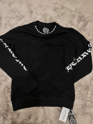 Svart Chrome Hearts x Comme des Garçons sweatshirt - Svart sweatshirt från Chrome Hearts x Comme des Garçons med vita tryck på ärmarna och nacken. Tröjan har ribbade muddar och rund hals. Materialet är mjuk bomull och passformen är normal. Snygga detaljer med logotyptryck både fram och bak.