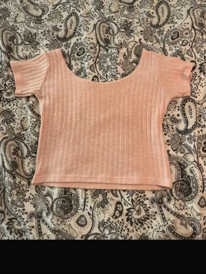 Ljusrosa ribbad croppad topp - Säljer en ljusrosa croppad topp med ribbad struktur och korta ärmar. Toppen har en bred, rundad urringning och är tillverkad i ett mjukt stretchigt material som sitter tajt mot kroppen. Perfekt till jeans eller kjol för en enkel men snygg look.