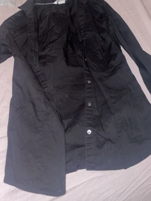 Svart skjorta från H&M, strl 34 - Svart skjorta från H&M i storlek 34. Klassisk modell med lång ärm, knappar framtill och vid manschetten. Skjortan är gjord i mjuk bomull och har en stilren look som funkar till många olika outfits.