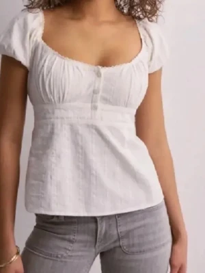 Vit blus med puffärm och knappar - Säljer en vit blus med korta puffärmar och v-ringad hals. Blusen har dekorativa knappar framtill och lätt rynkade detaljer vid bysten. Materialet är tunt och har en lätt glansig yta, vilket ger en fräsch och somrig känsla. Perfekt att styla med jeans eller kjol.