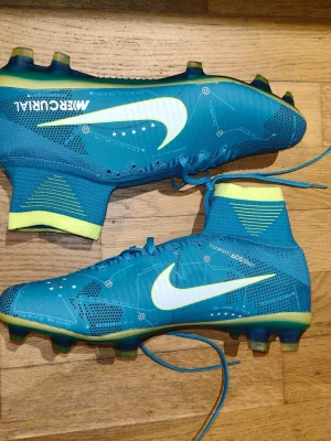 Nike Mercurial blå fotbollsskor - Säljer ett par Nike Mercurial fotbollsskor i blått med neongula detaljer och vit swoosh. Skorna har snörning, hög strumpkrage och mönstrad ovandel i syntetmaterial. Perfekta för dig som vill ha grym touch och stil på planen.