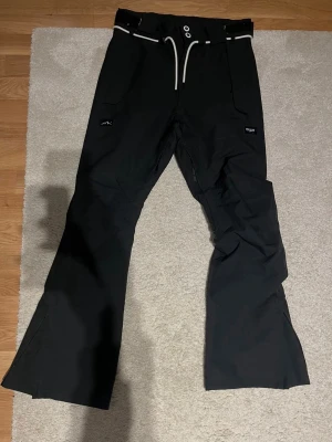 Svarta täckbyxor från Dope XXS - Svarta täckbyxor från Dope med vita snörningsdetaljer i midjan och praktiska fickor både fram och bak. Byxorna har bootcut-snitt och är gjorda i slitstark polyester, perfekta för vintern och snowboarding. Snygg logga på bakfickan och justerbar midja. Perfekt för vintersäsongen!