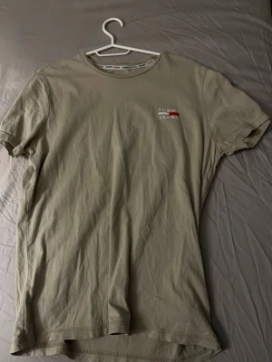 Beige t-shirt från Tommy Jeans - Säljer en beige t-shirt från Tommy Jeans i storlek M. T-shirten har rund hals, korta ärmar och en liten logga på bröstet. Materialet är mjuk bomull och passformen är normal. Perfekt för dig som gillar stilrena och enkla plagg med diskret branding.