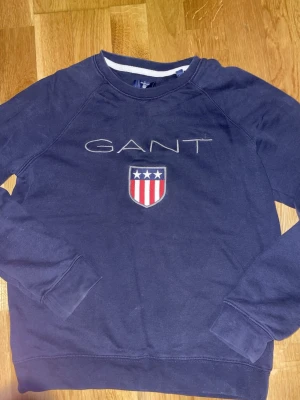 Gant hoodie - Säljer en marinblå sweatshirt från GANT med broderad logga och sköld med stjärnor och ränder på bröstet. Tröjan har rund halsringning och långa ärmar. Perfekt för dig som gillar klassisk och sportig stil.