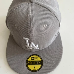 Grå New Era 59FIFTY LA keps - Snygg grå 59FIFTY keps från New Era med LA-logga broderad framtill i vitt. Klassisk flat peak och strukturerad passform. Kepsen är tillverkad i bomull och polyester, perfekt för dig som gillar streetstyle och sportig look.