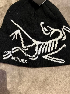 Svart Arc'teryx mössa med logga - Säljer en svart mössa från Arc'teryx med deras ikoniska vita fossil-logga stickad över hela framsidan. Mössan är tillverkad i mjukt syntetmaterial och har en clean, modern look som passar perfekt till kalla dagar.