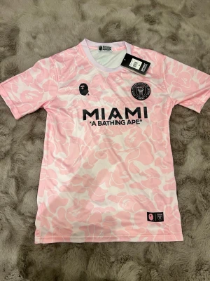 Miami Bape Jersey - Bilderna visar en rosa och vit fotbollströja från A Bathing Ape x Inter Miami, med camo-mönster och tryck. Ingen sko syns på bilderna.