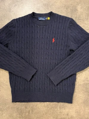 Mörkblå stickad tröja Polo Ralph Lauren - Kabelstickad mörkblå tröja från Polo Ralph Lauren med klassisk röd logga broderad på bröstet. Rund halsringning och långa ärmar. Perfekt för dig som gillar tidlös och stilren design.