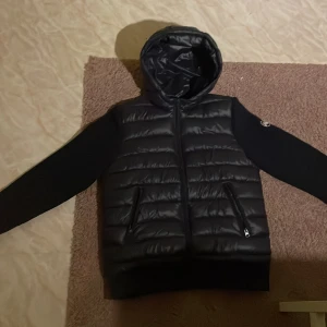 Svart pufferjacka från Maggiore - Svart pufferjacka från Maggiore med huva och quiltad framsida. Jackan har långa ärmar i ett annat material än kroppen, två fickor med dragkedja och en snygg logga på ärmen. Perfekt för dig som gillar stilren och sportig look. Andvänd bara 3 gånger 