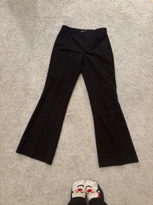 Svarta bootcut-byxor Gina Tricot - Svarta bootcut-byxor från Gina Tricot i storlek 34. Byxorna har klassisk midja, bälteshällor och är tillverkade i ett mjukt bomullsmaterial med lite stretch. Snyggt utsvängda ben som ger en trendig siluett. Perfekta att styla med sneakers eller chunky boots.