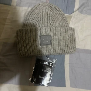 Acne studios mössa - Snygg grå ribbstickad mössa från Acne Studios med ikonisk ansiktslogga framtill. Tillverkad i 100% ull för extra värme och stil. Perfekt för dig som vill ha en clean och trendig look under kalla dagar.