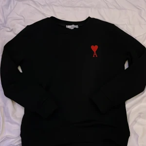 Svart sweatshirt från Ami Paris - Pris diskuteras gärna. Svart sweatshirt från Ami Paris med rött broderat hjärta och bokstaven A på bröstet. Klassisk rund halsringning och långa ärmar. Tillverkad i mjuk bomull för en skön och avslappnad vibe. 