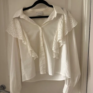 Vit volangskjorta från Neo Noir - Superfin vit skjorta från Neo Noir med stora broderade volanger över axlarna och fram. Skjortan har klassisk krage, knäppning framtill och långa ärmar med breda manschetter. Perfekt för dig som gillar romantiska detaljer och en trendig look.