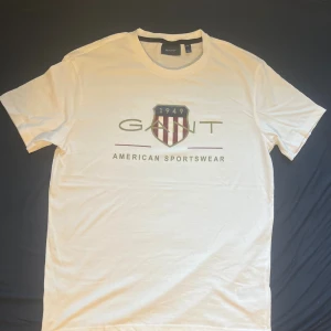 Vit GANT t-shirt med tryck - Vit t-shirt från GANT i bomull med klassiskt tryck framtill med sköld och texten 'American Sportswear'. Rund halsringning och korta ärmar. Perfekt för en clean och sportig look.