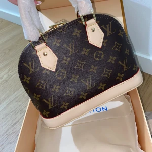 Louis Vuitton väska  - Säljer en ikonisk handväska från Louis Vuitton i klassiskt monogrammönster. Väskan är brun med beiga detaljer och guldfärgade metalldelar. Tillverkad i skinn med rundad form och två handtag. Perfekt för dig som vill ha en lyxig och tidlös accessoar❤️