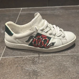 Gucci ace sneakers - Säljer ett par vita Gucci sneakers i skinn med broderad röd och svart orm på sidan, klassiska gröna och röda ränder samt metallicröd och metallicgrön häl. Skorna har snörning och rund tå, och är tillverkade i Italien. Perfekt för dig som vill sticka ut med lyxig streetstyle.