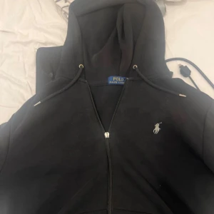 Svart hoodie från Polo Ralph Lauren - Svart hoodie från Polo Ralph Lauren med dragkedja framtill och klassisk huva med snörning. Tröjan har det ikoniska broderade Polo-logot på bröstet och är tillverkad i mjuk bomull. Perfekt för en chill och stilren look.