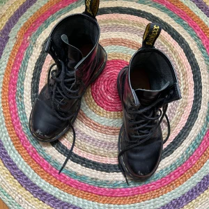 Svarta Dr. Martens boots i skinn - Klassiska svarta Dr. Martens boots i skinn med rund tå, snörning och den ikoniska gula sömmen runt sulan. Grov, mönstrad sula för extra grepp och slitstyrka. Perfekta för dig som vill ha en tidlös och edgy stil.