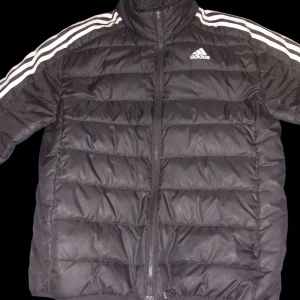 Svart pufferjacka Adidas - Svart pufferjacka från Adidas med klassiska vita ränder längs axlarna och ärmarna. Jackan har hög krage, dragkedja framtill och Adidas-logga på bröstet. Perfekt för kyliga dagar och har en sportig vibe.