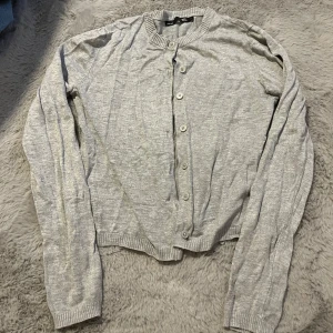 Grå kofta från 157 med knappar - Ljusgrå kofta från 157 i storlek M. Den har långa ärmar, ribbade muddar och knäppning framtill med flera knappar. Perfekt att slänga över en t-shirt eller linne när det blir lite kyligt.