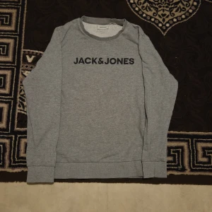 Grå sweatshirt från Jack & Jones XL - Säljer en stilren grå sweatshirt från Jack & Jones i storlek XL, passar även L och M. Tröjan har svart logotyptryck på bröstet, rund halsringning och långa ärmar. Tillverkad i mjuk bomullsmix som är skön att ha på sig. Perfekt för en chill och enkel look. Pris kan diskuteras.