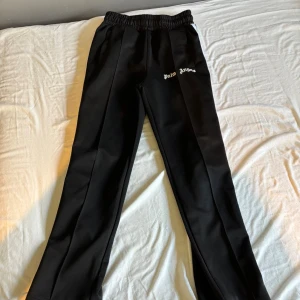 Palm Angels svarta mjukisbyxor - Svarta mjukisbyxor från Palm Angels med vit logotyp på framsidan och vita ränder längs sidorna. Byxorna har elastisk midja och raka ben för en chill och snygg look. Perfekta för streetwear och avslappnade dagar.