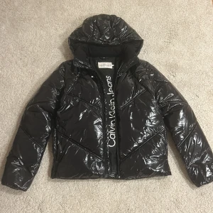 Svart pufferjacka från Calvin Klein Jeans - Snygg svart pufferjacka från Calvin Klein Jeans med glansig finish och huva. Jackan har dragkedja framtill, diskret logga på ärmen och coolt mönstrade sömmar. Perfekt för kalla dagar när du vill hålla stilen. Passar som Xs/S. Priset kan diskuteras!