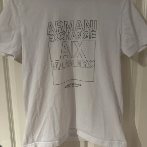 Vit Armani Exchange t-shirt med tryck - Vit t-shirt från Armani Exchange i mjuk bomull. Framsidan har ett stilrent svart tryck med texten 'ARMANI EXCHANGE A|X MILAN NYC'. Klassisk rund hals och korta ärmar. Perfekt för dig som gillar enkel och snygg streetstil.