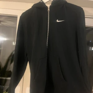 Svart Nike hoodie med dragkedja - Svart hoodie från Nike med vit logga på bröstet. Tröjan har dragkedja framtill, huva med snörning och två stora fickor. Perfekt för en avslappnad stil och funkar året runt. Klassisk och enkel design som alltid är rätt.