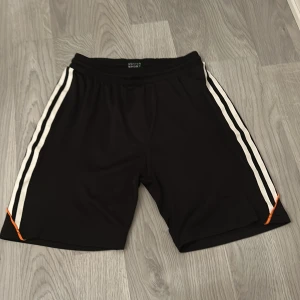 Svarta träningsshorts Active Sport - Svarta träningsshorts från Active Sport med vita ränder längs sidorna och orange detaljer. De har elastisk midja och är gjorda i lätt polyester som andas. Perfekta för sport eller gym, med loose passform och snygg kontrast mellan färgerna.