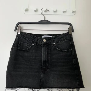 Svart jeanskjol från Pull&Bear - Svart jeanskjol från Pull&Bear med rå kant nertill och klassiska fem fickor. Kjolen har bälteshällor, knapp och dragkedja framtill. Snygg, kort modell som ger en cool vibe och passar perfekt till sneakers eller boots.