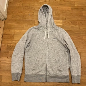 Ljusgrå hoodie från Hollister - Ljusgrå hoodie från Hollister med dragkedja, huva och snörning. Mjuk insida, två fickor framtill och broderad logga på bröstet. Perfekt för chill dagar och enkel att matcha med jeans eller joggers.