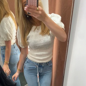 Vit blus med puffärm - Säljer en vit blus med puffiga korta ärmar och smockad struktur. Toppen har rund hals och är figurnära med en lätt volang vid ärmslut. Perfekt för dig som gillar en feminin och trendig stil.