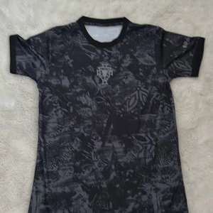 Svart mönstrad fotbollströja Portugal  - Unik svart fotbollströja från Portugal med grått abstrakt mönster över hela tyget. Tröjan har korta ärmar, rund hals och broderade detaljer på bröstet och ärmen. Materialet är lätt och andas, perfekt för träning eller match.