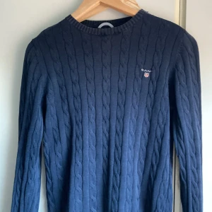 Marinblå kabelstickad tröja GANT - Snygg marinblå kabelstickad tröja från GANT med rund hals och broderad logga på bröstet. Tröjan har lång ärm och klassisk ribbad kant vid halsen. Perfekt för dig som gillar stilrena och tidlösa plagg med preppy vibe.