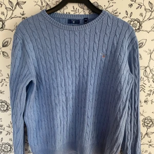 Ljusblå kabelstickad tröja GANT - Snygg ljusblå kabelstickad tröja från GANT med rund hals och broderad logga på bröstet. Tröjan har långärmad passform och ribbade muddar vid hals, ärmslut och nederkant. Perfekt för dig som gillar klassisk stil och mjuka material.