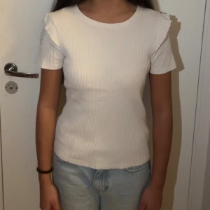 Vit ribbad topp med volang från Zara - Supersöt vit ribbad topp från Zara med korta ärmar och volangdetaljer vid axlarna. Toppen har en rund halsringning och är figurnära i modellen. Perfekt att styla med jeans eller kjol för en fräsch look.                                                                                Storlek M men funkar på Xs S 