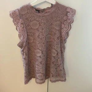 Rosa spetsblus från bubbleroom XS - Supersöt ljusrosa blus från bubbelroom i storlek XS. Blusen är ärmlös med vackra volangdetaljer längs axlarna och har ett heltäckande blommigt spetsmönster. Rund halsringning med knapp i nacken och fodrad kropp. Perfekt för dig som gillar romantisk stil!