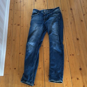 Blå jeans från Jack & Jones - Säljer ett par klassiska blå jeans från Jack & Jones med snyggt slitna detaljer och femficksmodell. Jeansen har normal passform och raka ben, perfekt för en avslappnad stil. Materialet är mjukt denim i bomull och färgen är mellanblå med ljusare partier.