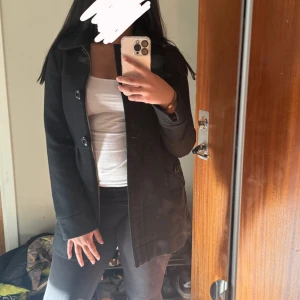 Svart kappa med knappar, stl 36 - Stilren svart kappa med stora knappar och dragkedja. Jackan har klassisk krage, markerad midja och fickor med lock och knappdetalj. Perfekt för dig som vill ha en enkel men snygg look till höst och vinter.