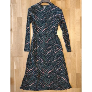 Grön långärmad klänning från Lindex - Snygg långklänning från Lindex i storlek M med zebramönster i grönt, svart och rosa. Klänningen har lång ärm, rund hals och är tillverkad i mjuk polyester. Passformen är normal och tyget har ett coolt mönster som sticker ut.