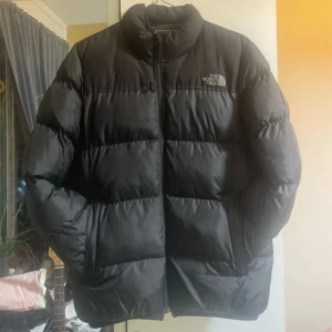 Svart pufferjacka från The North Face - Säljer en svart pufferjacka från The North Face med hög krage och dragkedja framtill. Jackan har logga på bröstet och ärmen, samt breda quiltade sektioner som ger en riktigt fet och puffig look. Perfekt för kalla dagar när du vill hålla stilen och värmen. Har ett litet hål på ena armen.