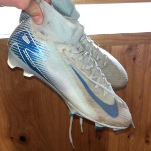 Nike Mercurial Zoom fotbollsskor blå/vit - Nike Mercurial Zoom fotbollsskor i ljusbeige och blått med högt skaft och snörning. Snyggt mönstrad med blå detaljer och Nike-logga på sidan. Skorna har platt sula med dobbar för gräsplan och är tillverkade i syntetmaterial för lätt känsla och bra grepp. Ingen påse.