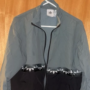 Grå och svart vindjacka från Adidas - Säljer en snygg vindjacka från Adidas i grått och svart med coolt band med vita trefoil-loggor runt hela jackan. Jackan har hel dragkedja framtill, hög krage och är tillverkad i lätt polyester. Perfekt för dig som gillar sportig och stilren streetwear.