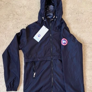  Canada Goose windbraker  - Snygg svart vindjacka från Canada Goose med huva och dragsko, klassisk logga på ärmen och hel dragkedja framtill. Jackan har en ficka med dragkedja och elastiska muddar vid ärmsluten. Perfekt för blåsiga dagar och enkel att matcha.