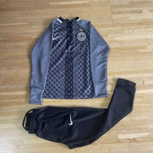 Grå långärmad PSG fotbollströja Nike - Snygg långärmad PSG fotbollströja från Nike i grått med svart geometriskt mönster framtill och klubbmärke på bröstet. Tröjan har Qatar Airways-tryck och är tillverkad i ett lätt, sportigt material som andas. Perfekt för träning eller match.