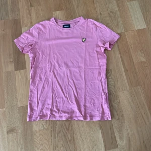 Rosa t-shirt från Lyle & Scott Junior - Snygg rosa t-shirt från Lyle & Scott Junior med klassisk rund hals och korta ärmar. På bröstet sitter den ikoniska broderade gula fågeln som ger en cool detalj. Mjuk och skön bomull som passar perfekt till jeans eller shorts.
