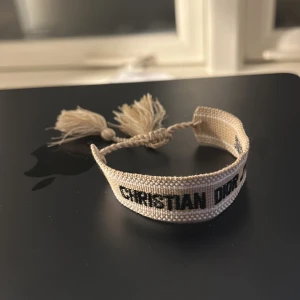 Beige Christian Dior J'Adior armband - Snyggt beige armband från Christian Dior med svart broderad text 'J'ADIOR'. Armbandet är gjort i ett vävt tygmaterial och har två tofsar i ändarna samt justerbar knytning. Perfekt accessoar för dig som gillar designer och vill sticka ut med något unikt.